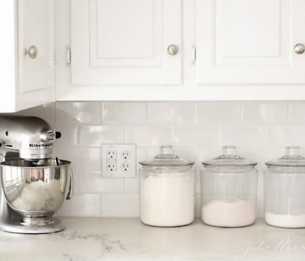 A white kitchen with marble countertops, a stand mixer and glass canisters of baking supplies.