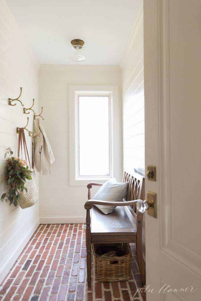 A white mudroom with horizontal paneling, brass hooks and a wooden bench.