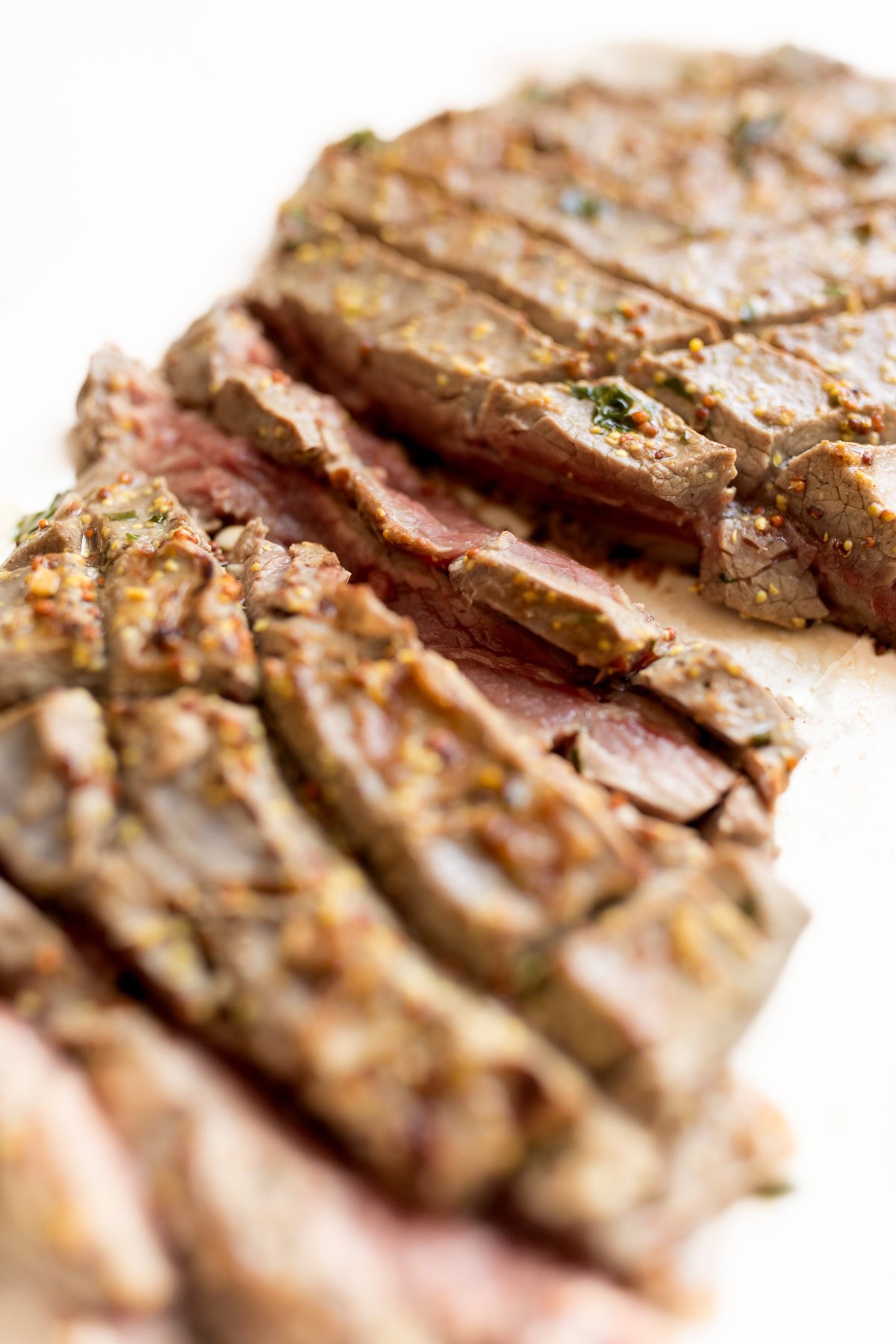 Close-up of sliced, broiled steak on a white surface, showcasing the meat's texture and seasoning. The steak appears to be medium-rare with visible herbs and spices.