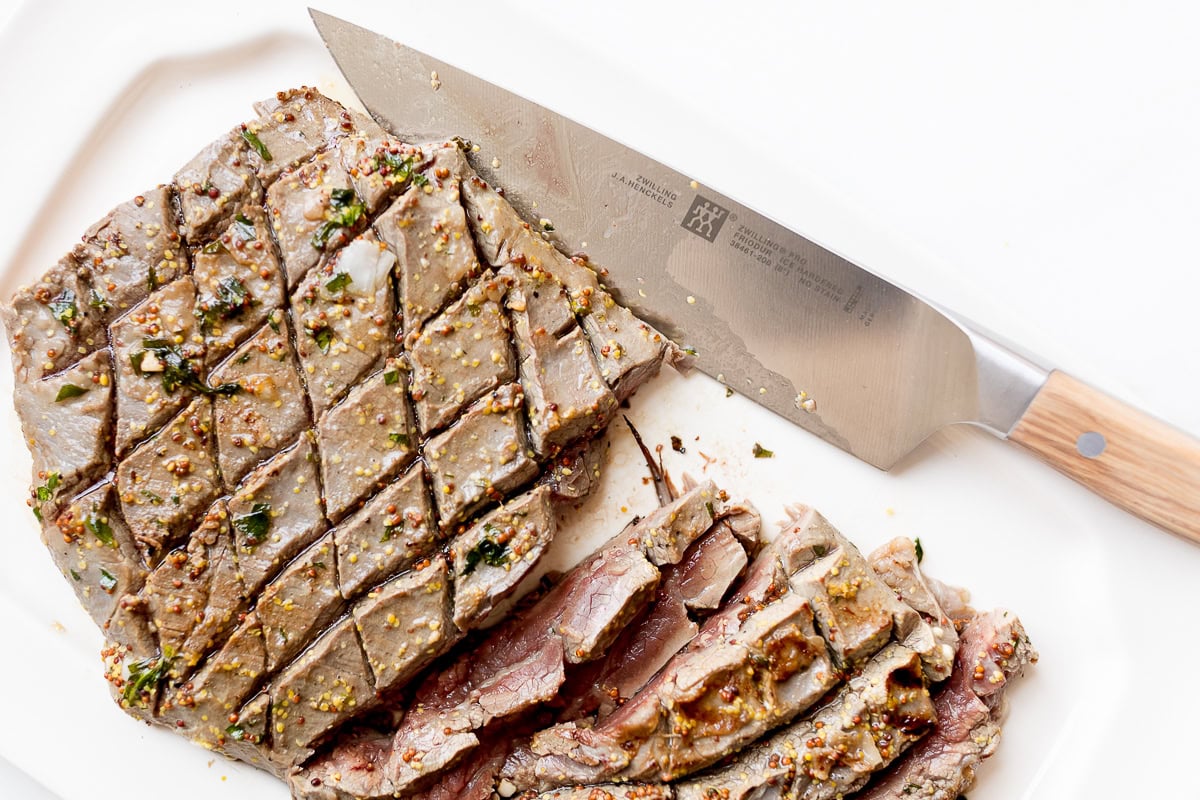 Sliced, seasoned, and broiled steak with a cross-hatched pattern on a white plate. A large chef's knife with a wooden handle rests beside the meat.