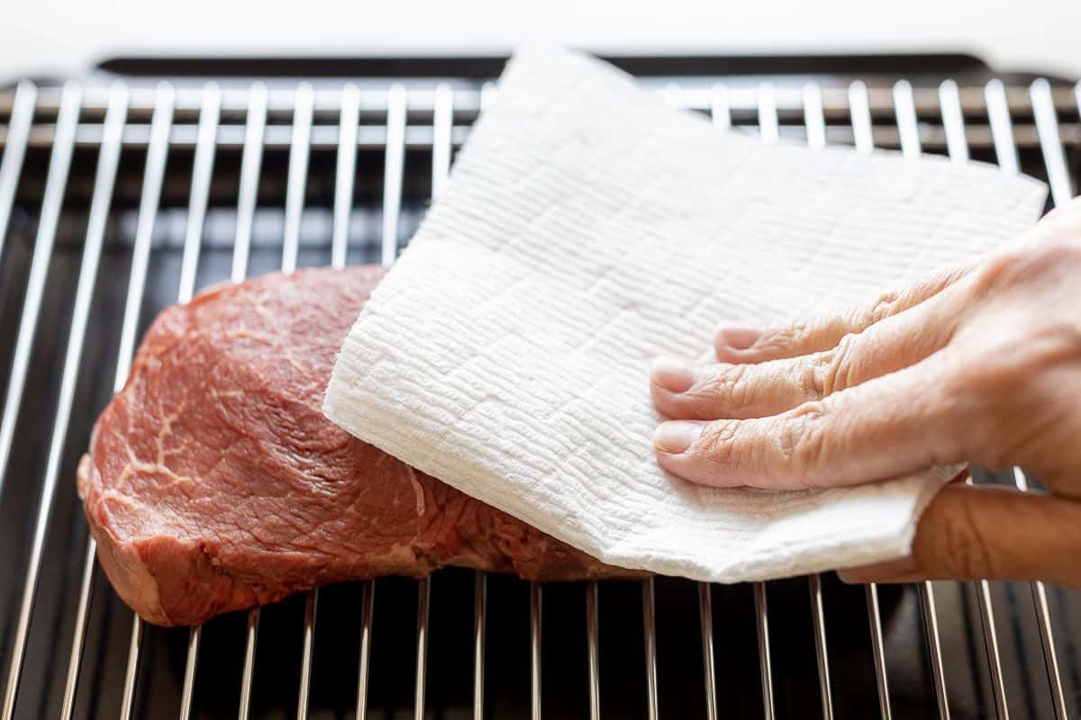 A hand using a paper towel to dry steak on a broiling rack.