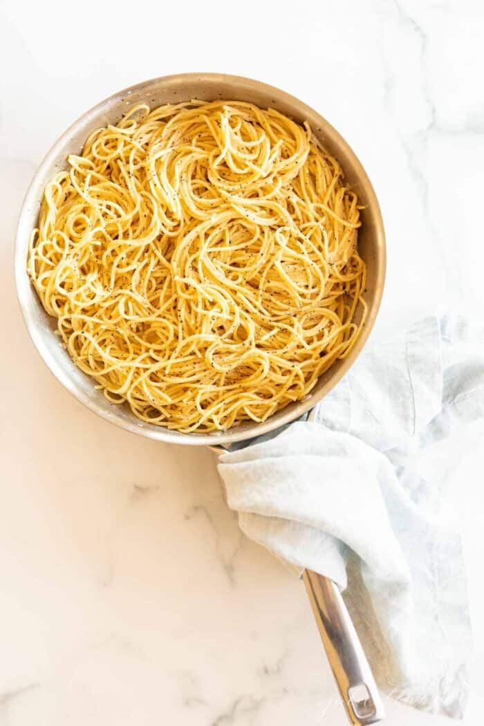 A silver pan full of cacio e pepe noodles, blue towel wrapped around handle on a marble surface.