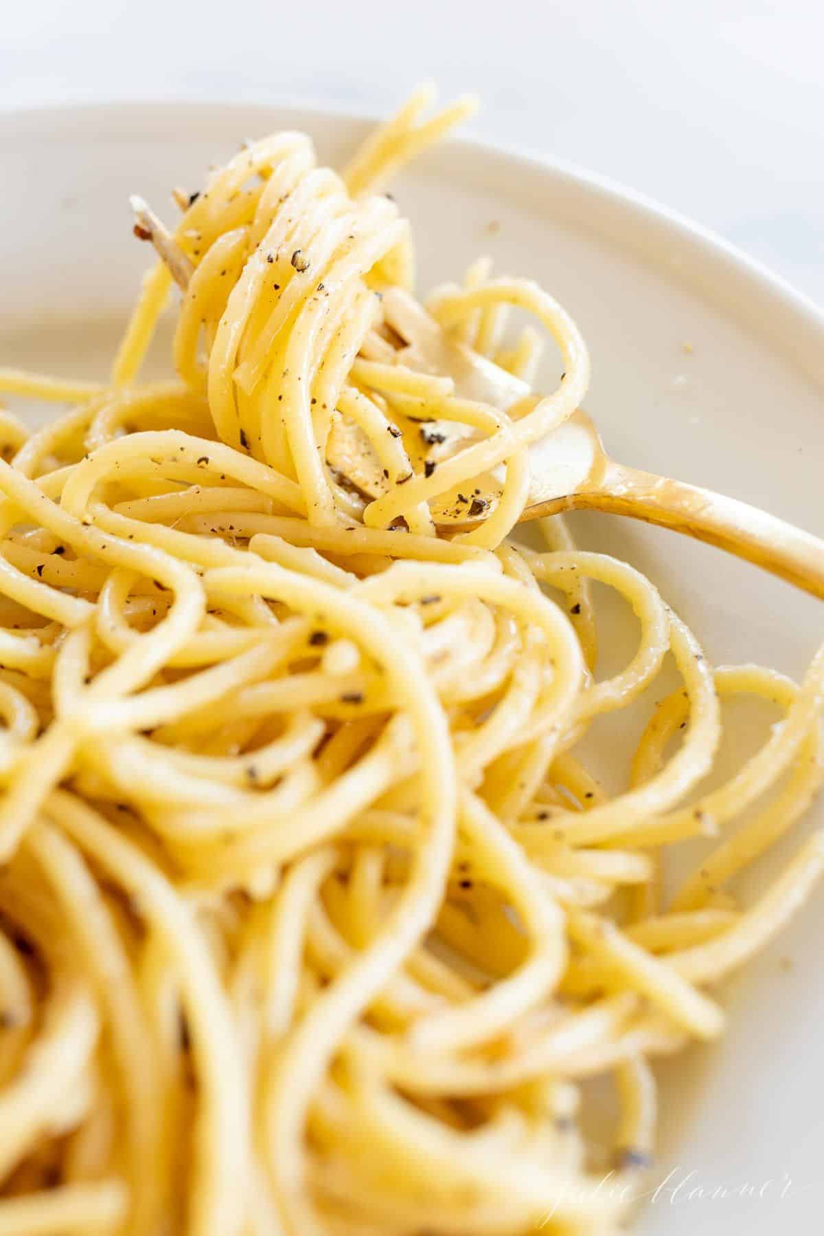 Image featuring a white plate full of cacio e pepe with spaghetti noodles, gold fork twirled with pasta on the side of plate.