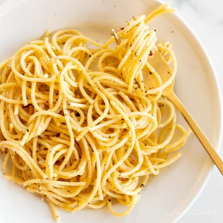 A square image featuring a white plate full of cacio e pepe with spaghetti noodles, gold fork twirled with pasta on the side of plate.