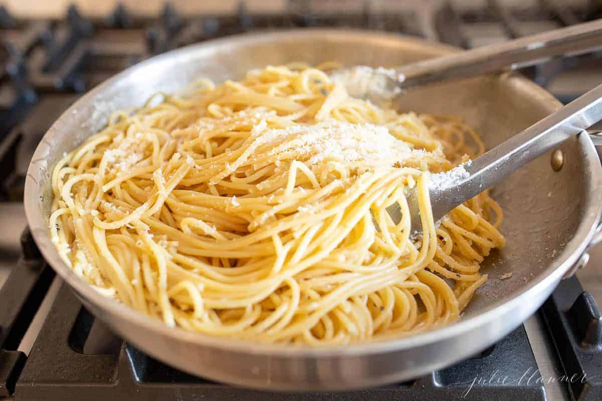 A silver pan on a stove top, filled with cooked spaghetti, silver tongs pulling it out of pan.