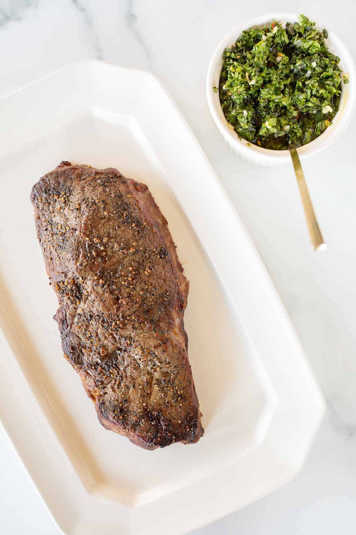 A steak on a white platter, bowl of chimichurri sauce to the side with a gold spoon.