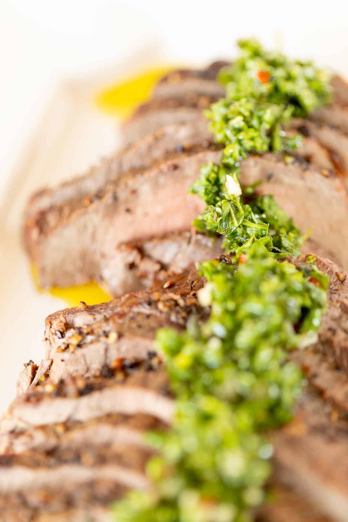 Sliced steak on a white platter, topped with chimichurri.