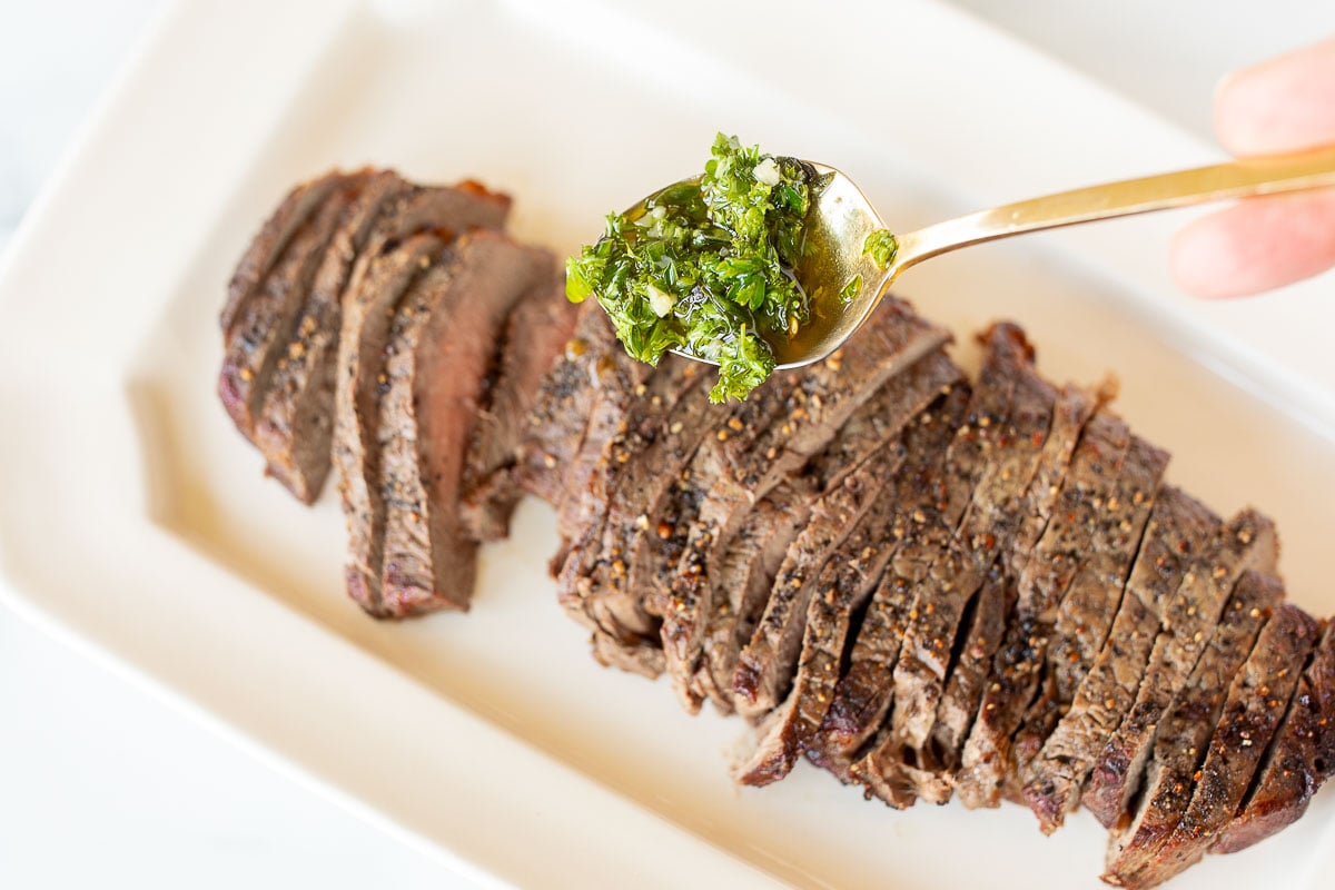 Sliced steak on a white plate, with a spoon full of a homemade chimichurri recipe coming in.