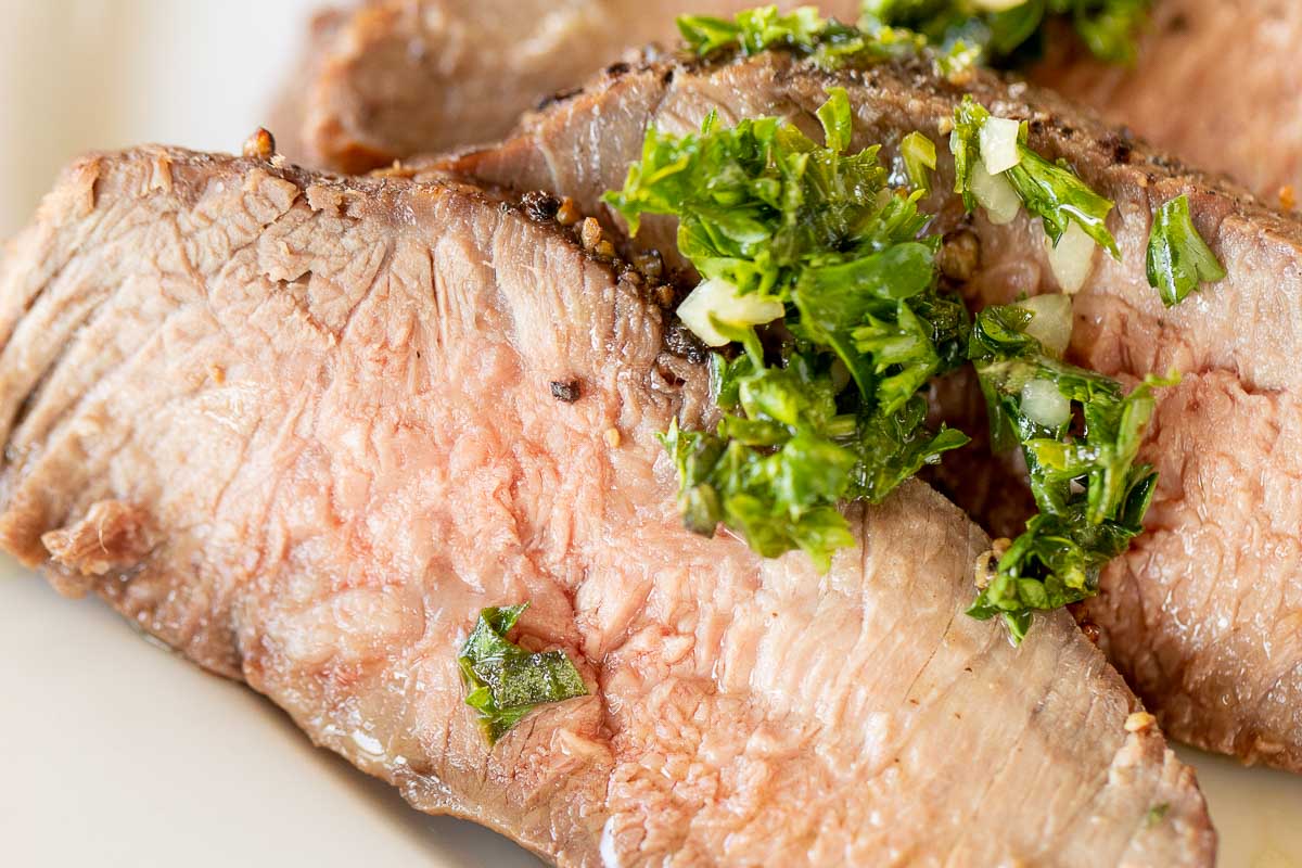 Sliced steak on a white plate, topped with chimichurri.
