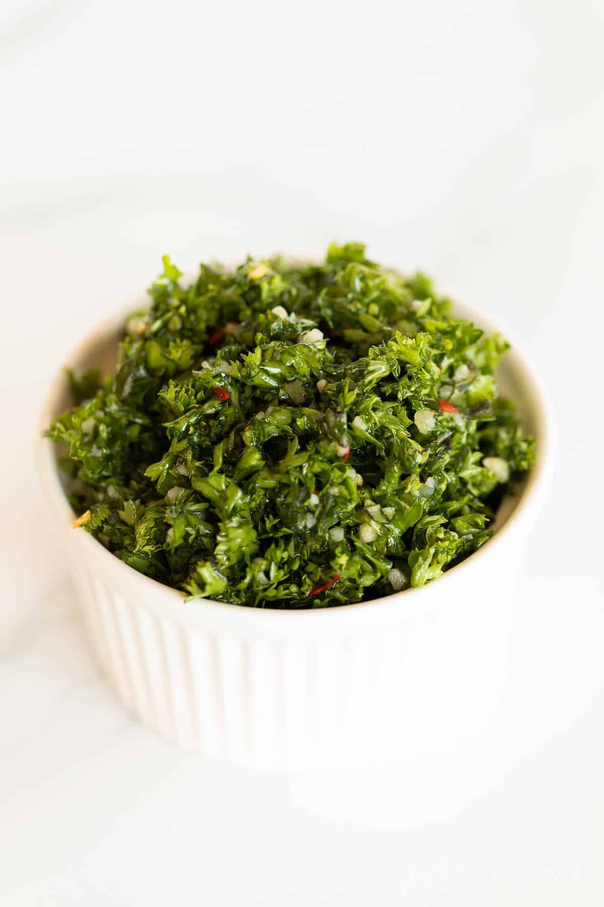 A white ramekin full of chimichurri on a marble surface.