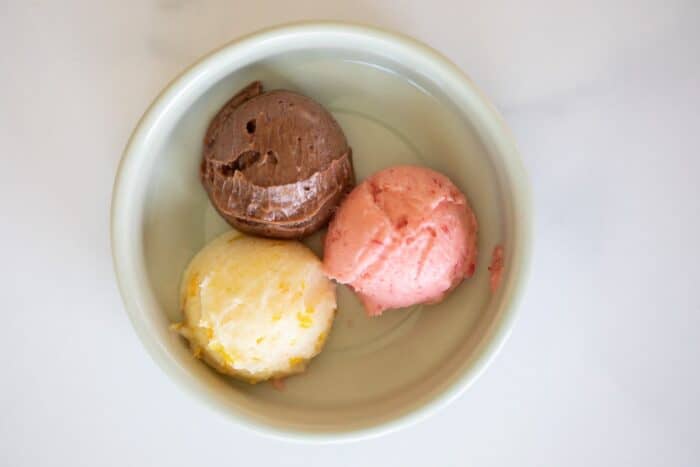 A white ramekin on a marble surface filled with three scoops of flavored butter, one chocolate, one strawberry and one orange.