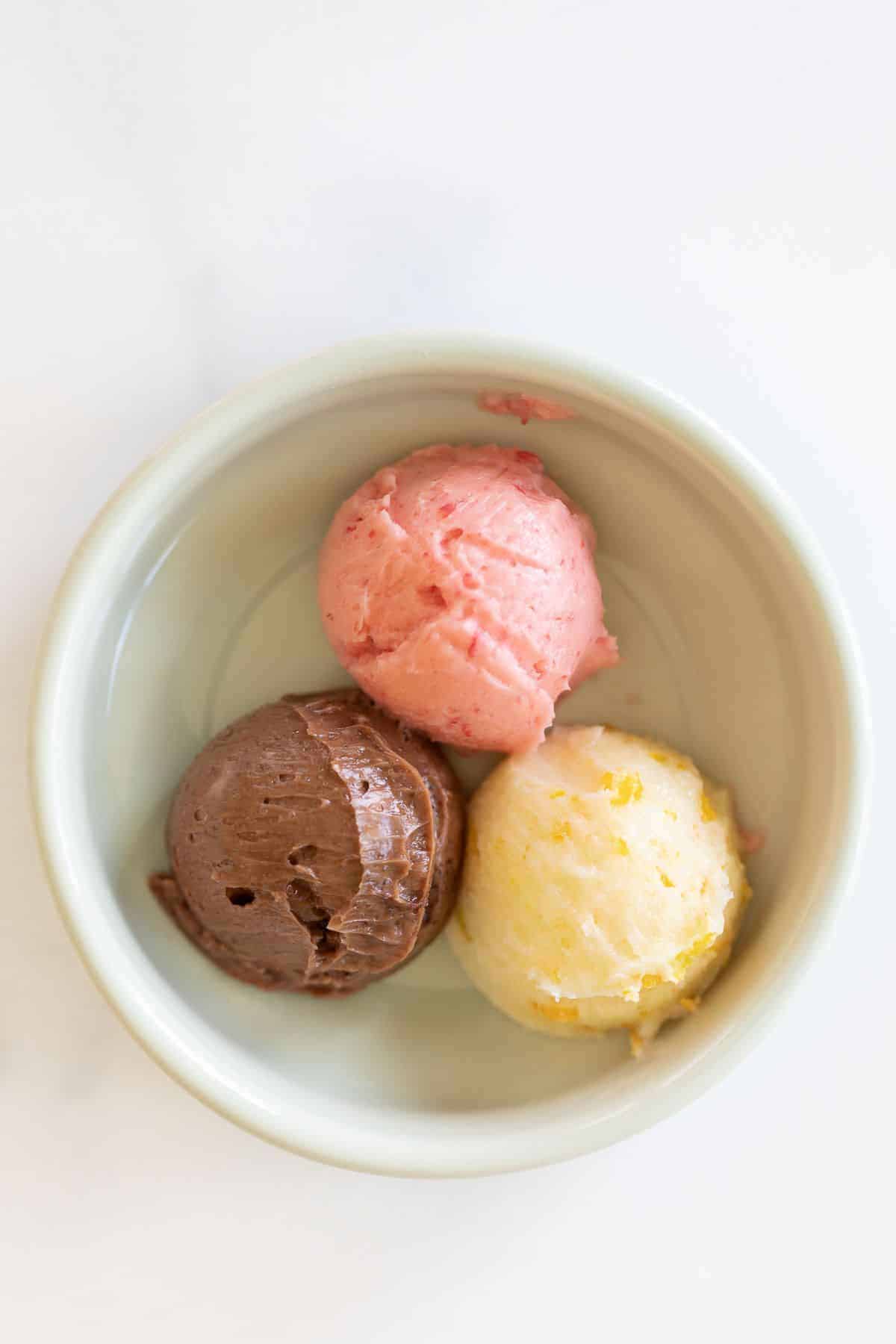 A white ramekin on a marble surface filled with three scoops of flavored butter, one chocolate, one strawberry and one orange.
