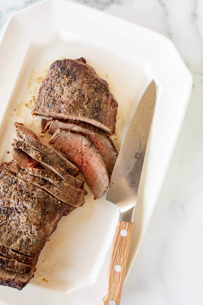 A broiled steak sliced on a white platter, large knife to the side.