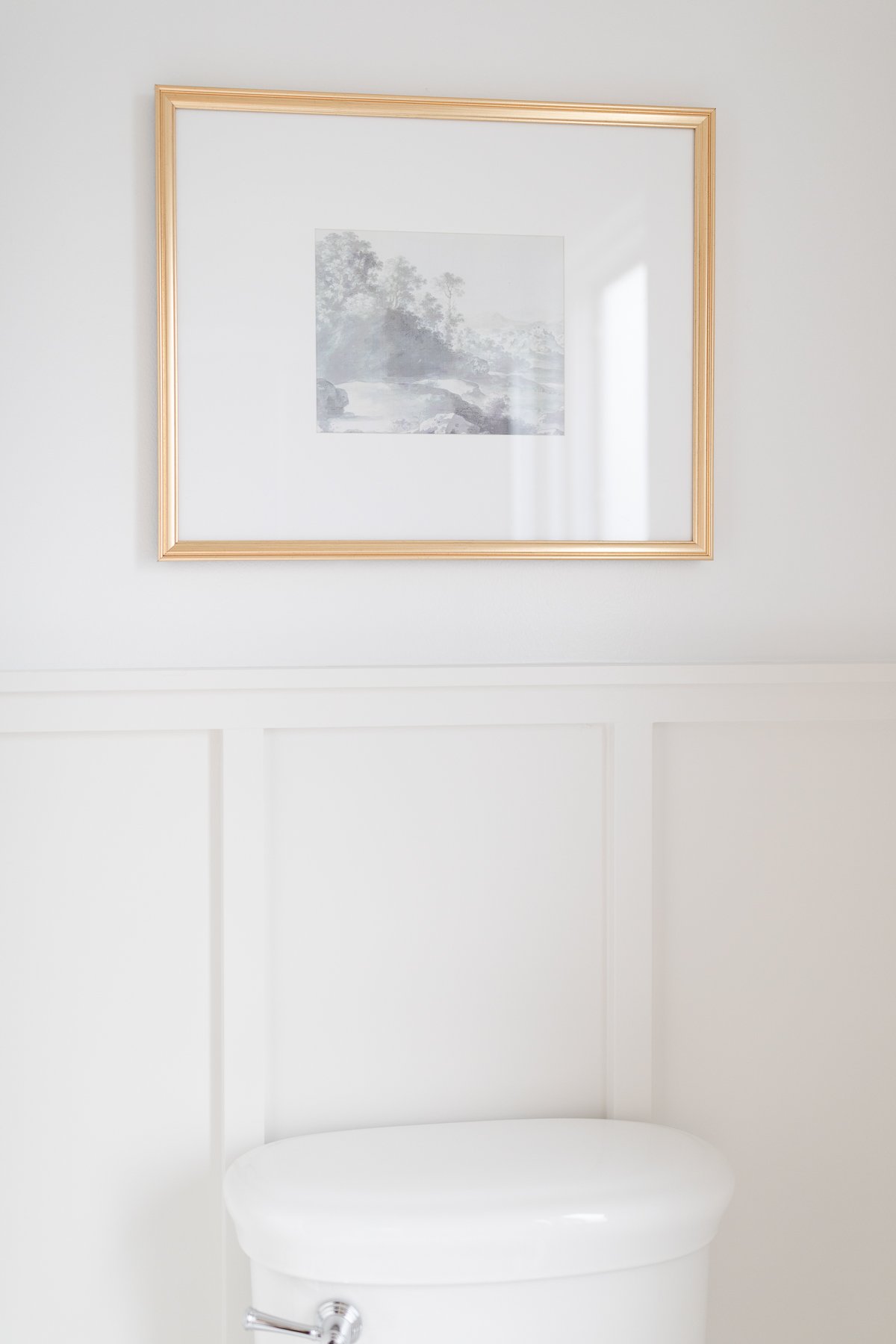 A toilet in a white bathroom with a framed picture sourced from Studio McGee above it.