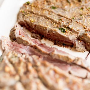 Close-up of a broiled steak on a white plate, showcasing a pink, juicy interior and a browned exterior with visible herbs and spices.