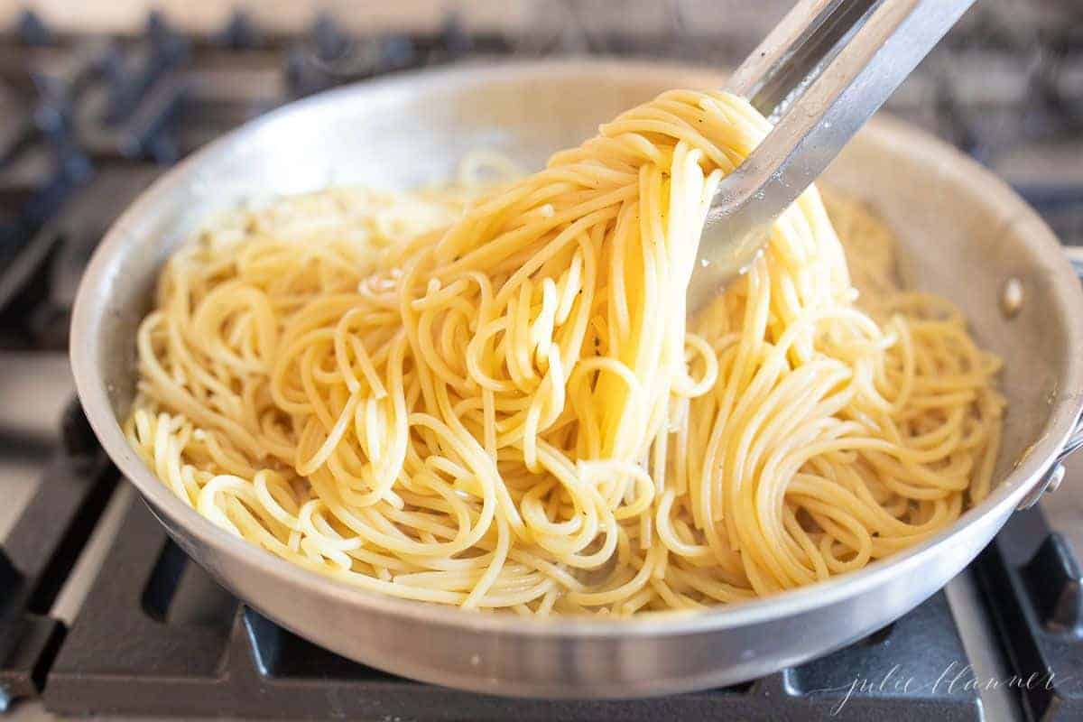 A silver pan on a stove top, filled with cooked spaghetti, silver tongs pulling it out of pan.