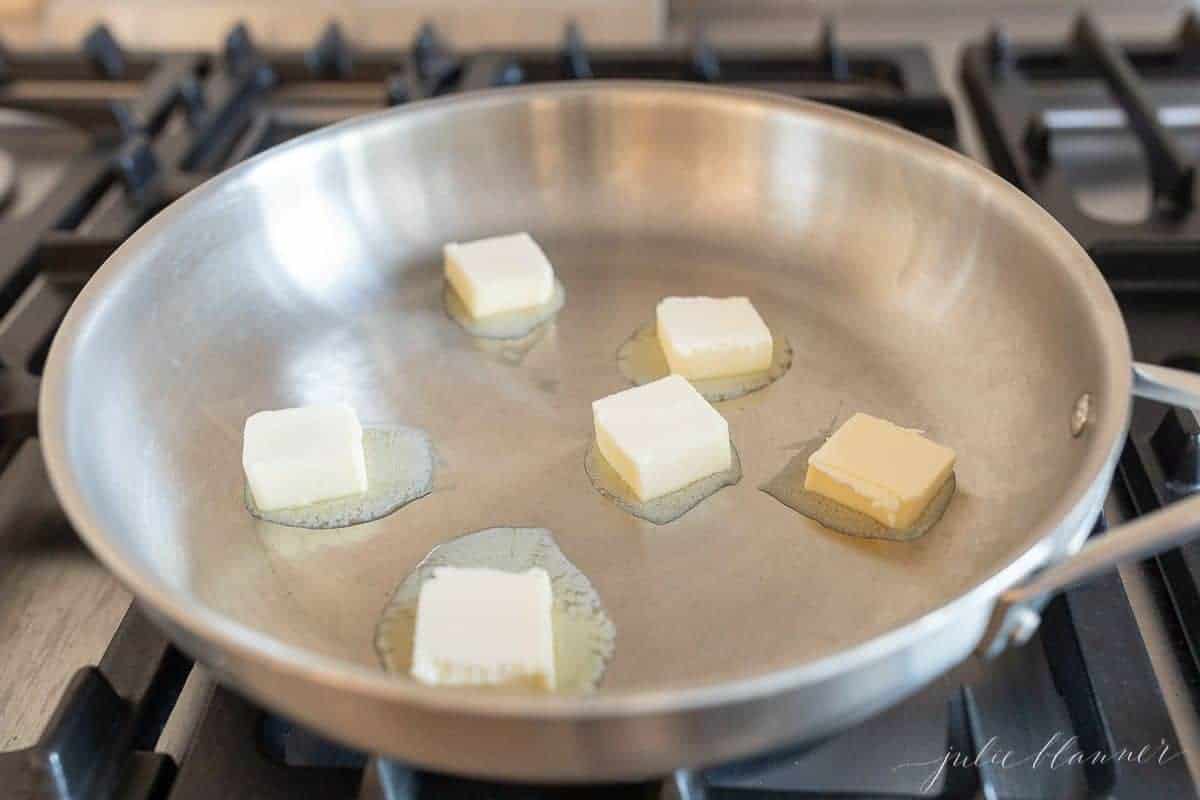 A silver pan on a stovetop, filled with melting pats of butter.