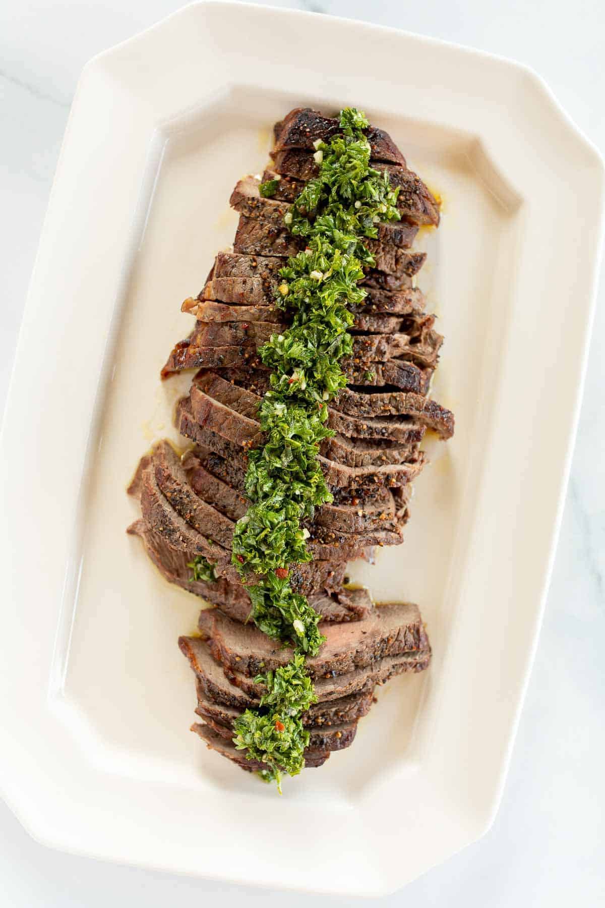 Sliced steak on a white platter, topped with chimichurri.
