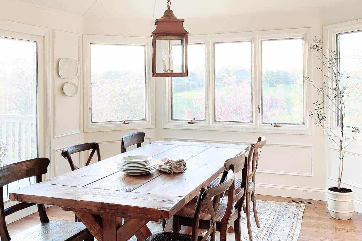 A white dining room with lots of windows, a wooden farm table and an indoor olive tree in the corner.