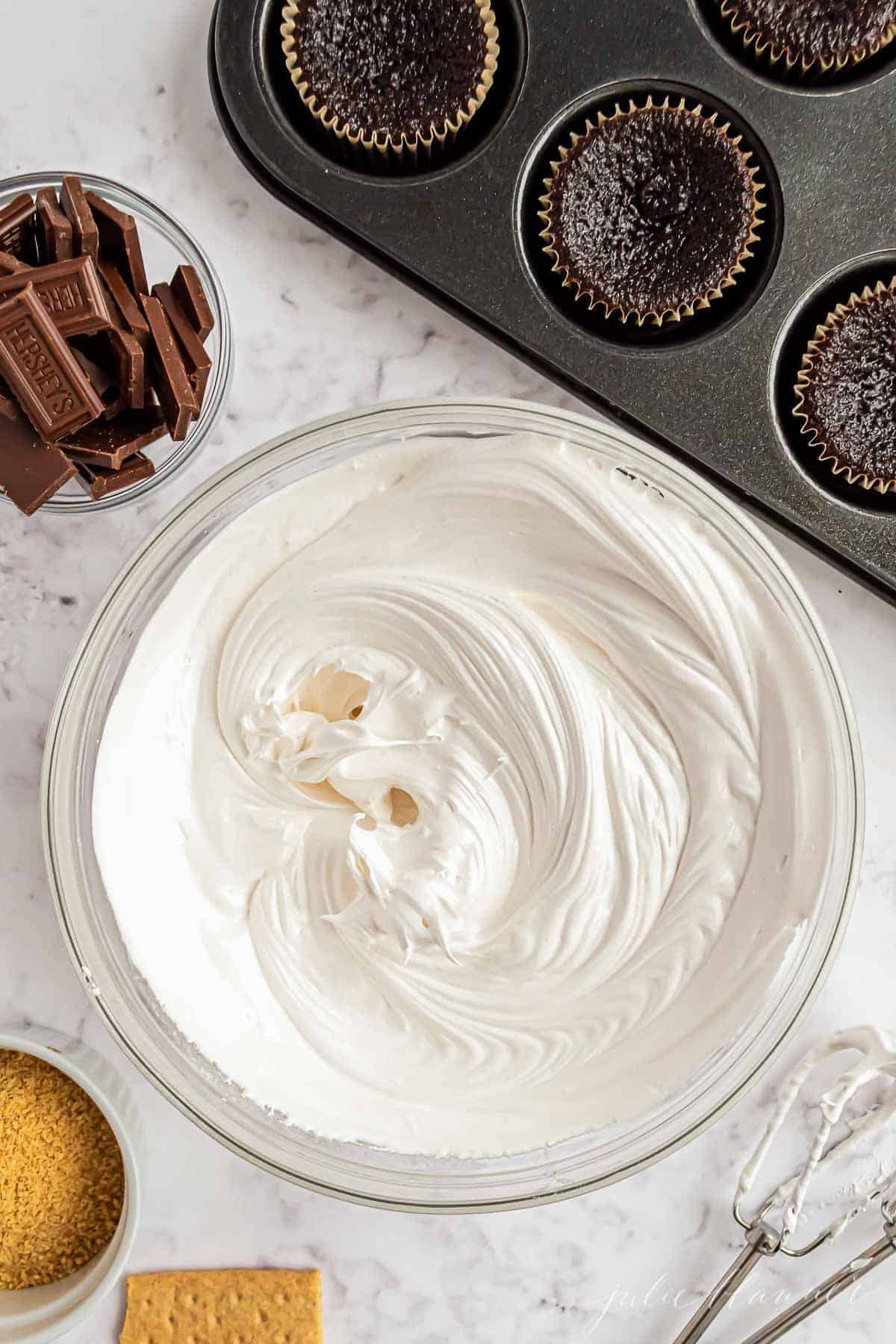 A clear glass bowl full of marshmallow icing, near chocolate cupcakes.