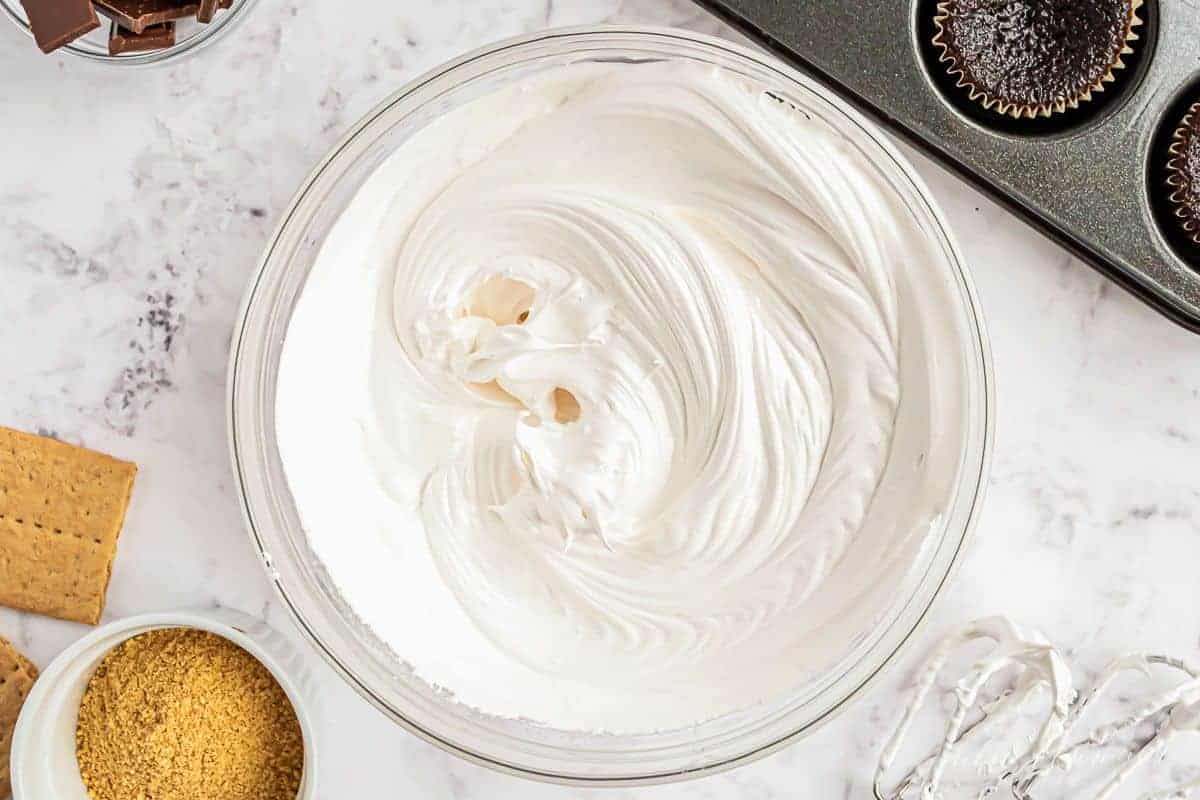 A clear glass bowl full of marshmallow icing, near chocolate cupcakes.
