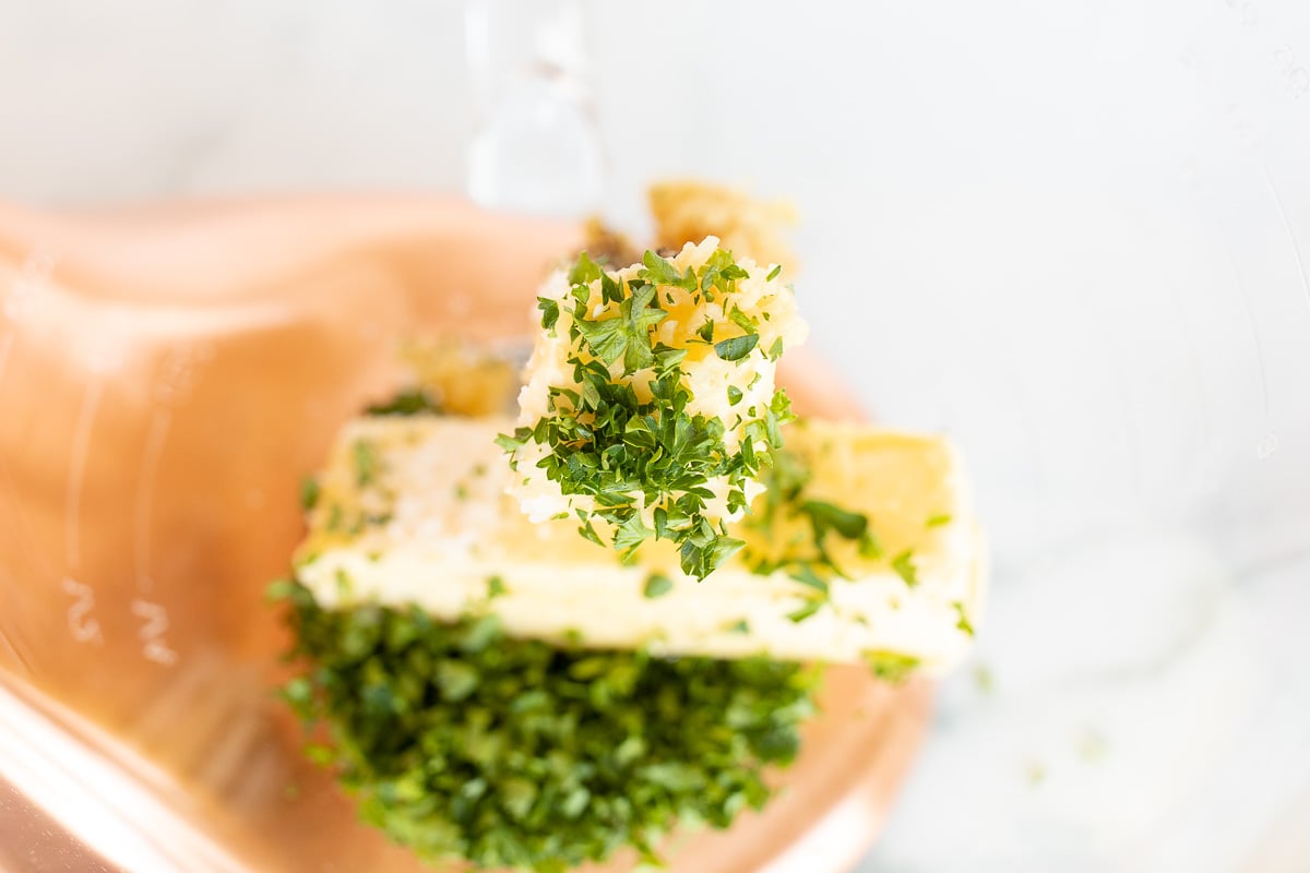 A stick of butter and fresh herbs inside the glass bowl of a mixer.