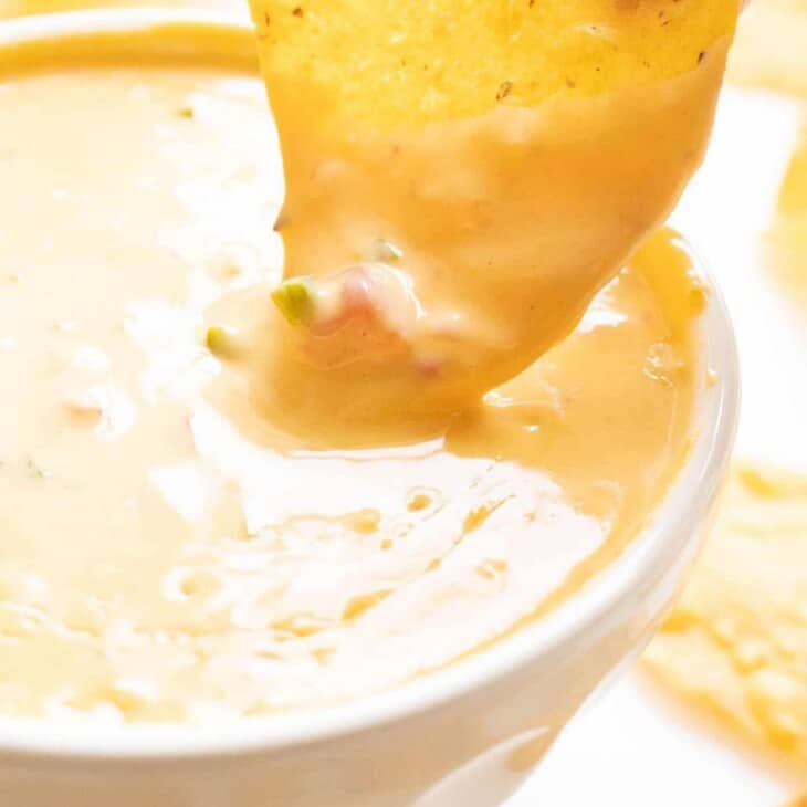 A chip is dipping into a white bowl of cheese dip.