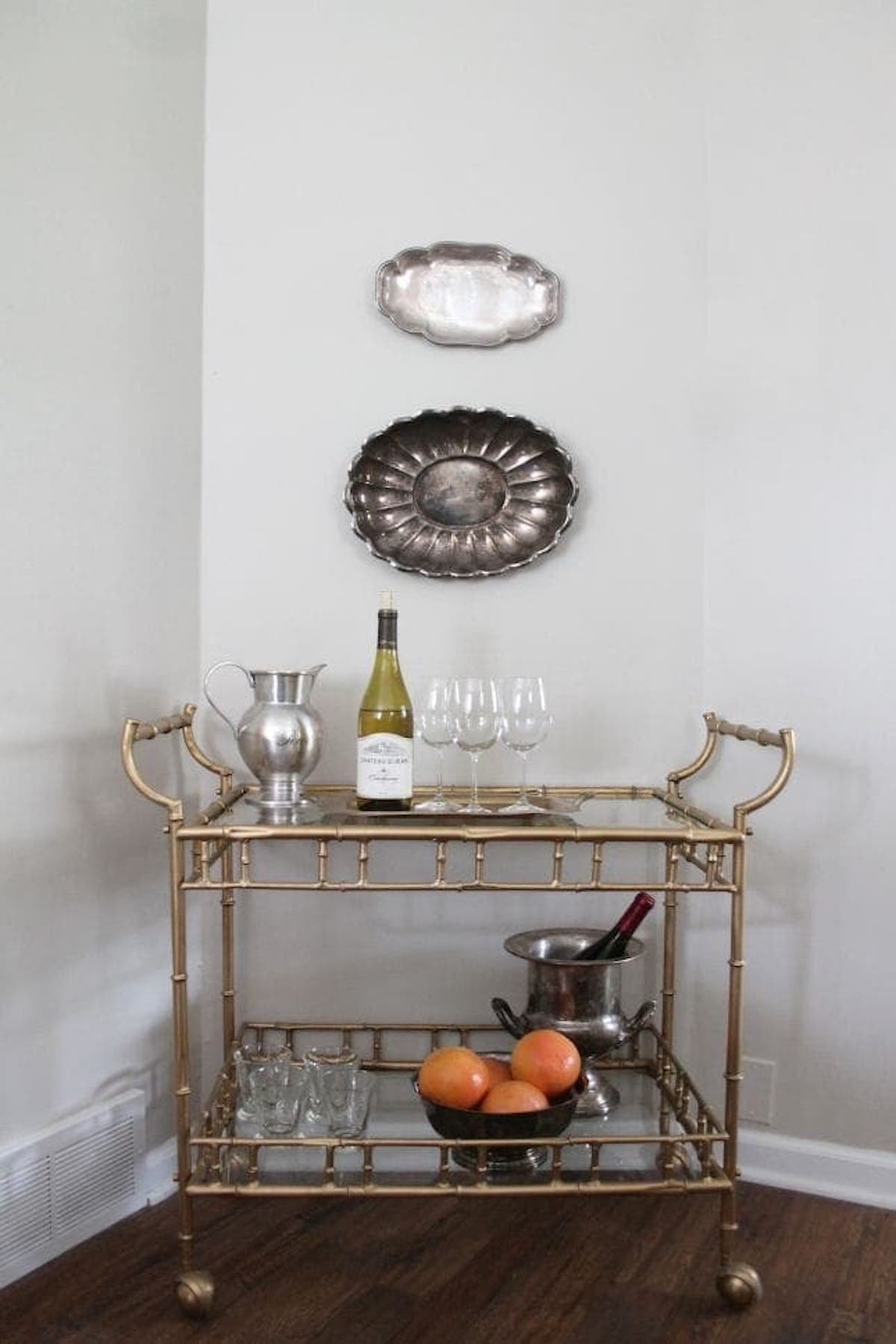 A brass bar cart with two shelves holds a wine bottle, four glasses, a pitcher, a bowl of oranges, and an ice bucket with a bottle inside. Two silver trays are mounted on the wall above it, beautifully contrasting with the Sherwin Williams Accessible Beige paint on the walls.
