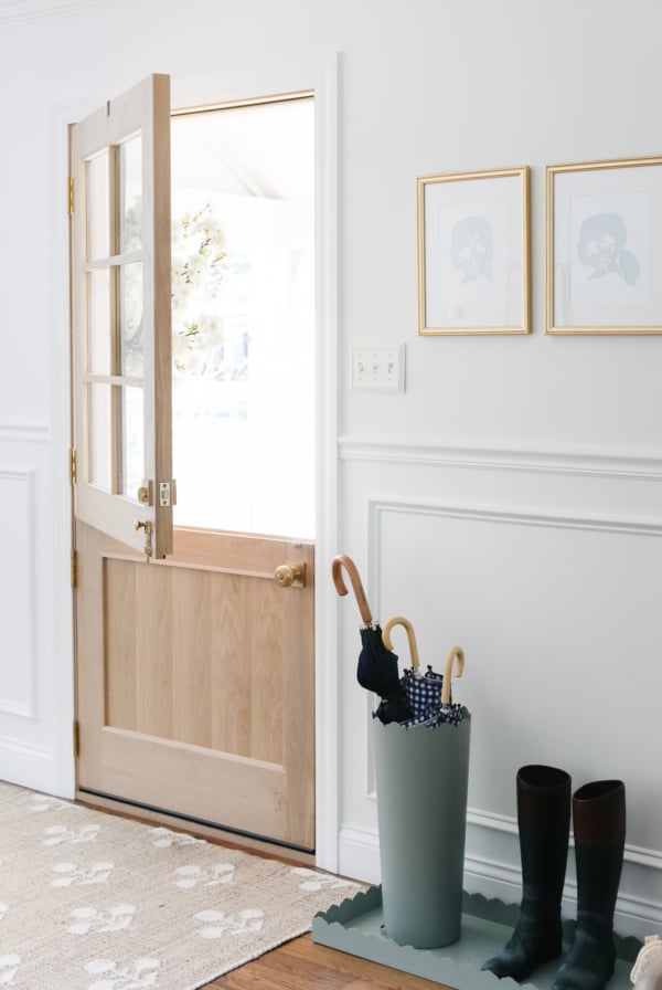 A bright interior entryway with an open wooden door, a pair of boots, and an umbrella in a stand, featuring the best paint for trim on the framed artwork on the wall.