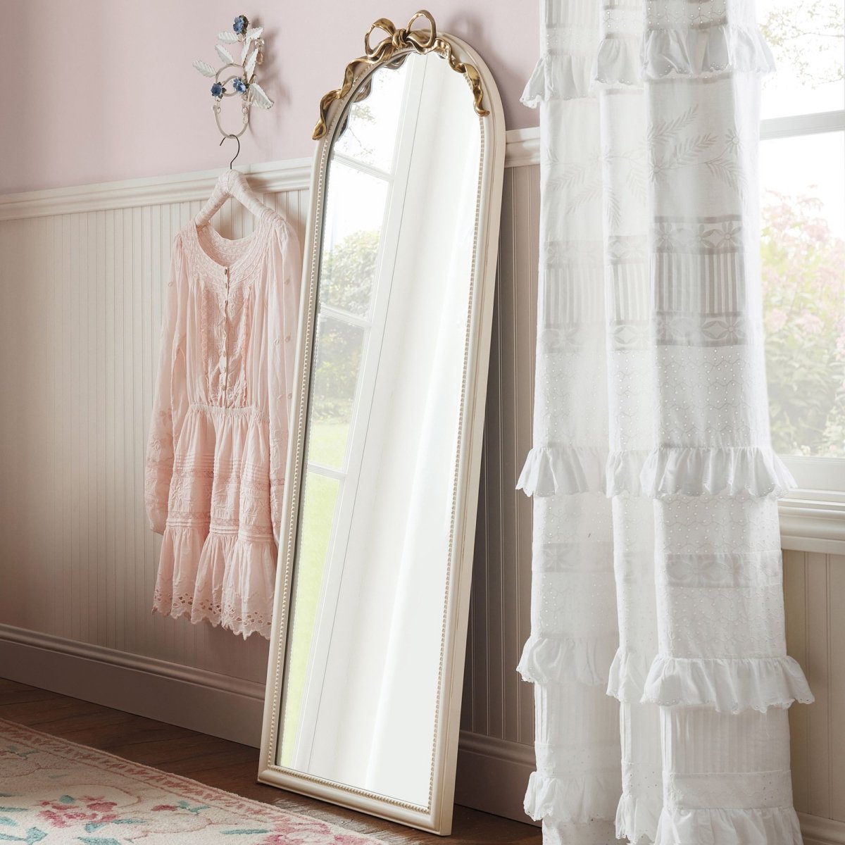 A coquette mirror hangs in a pink room with curtains and a dresser, perfect for tween bedroom ideas.