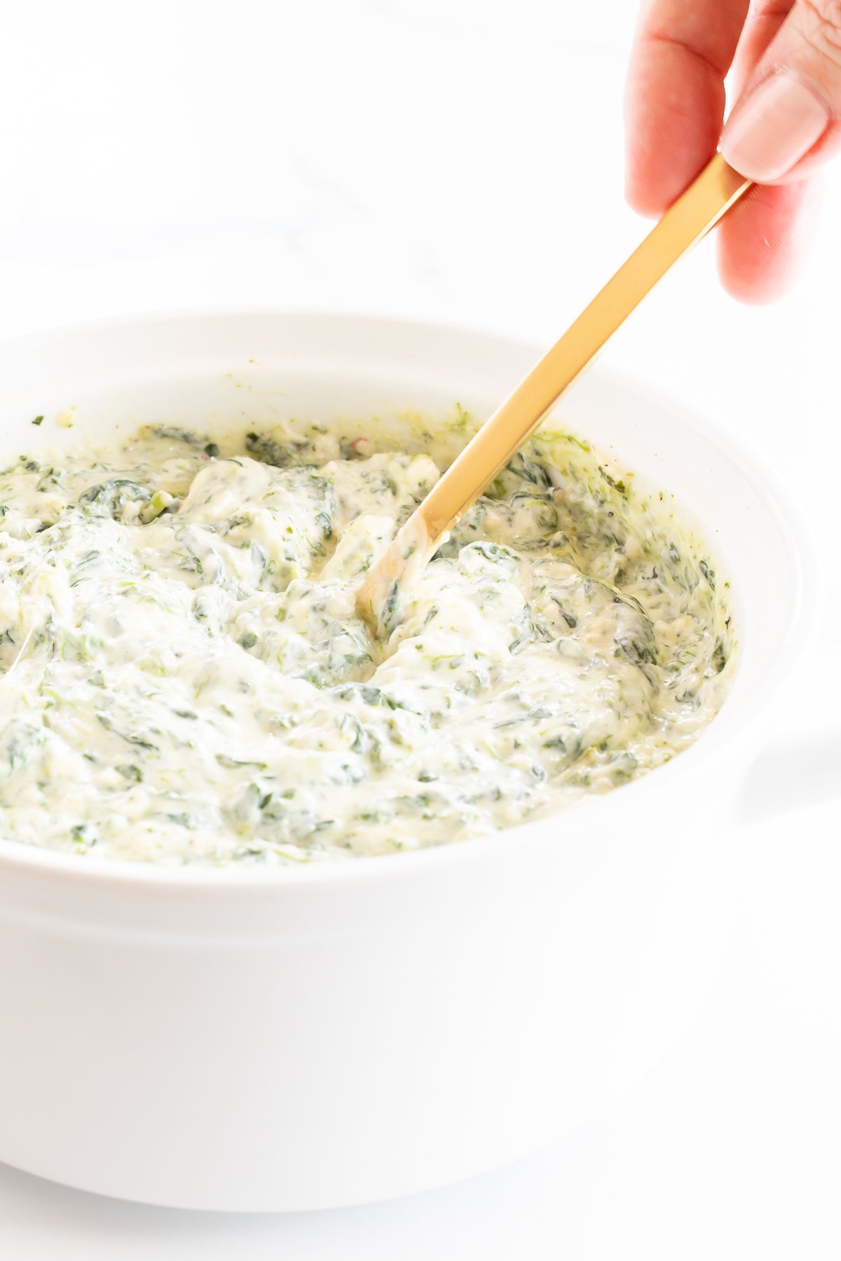 A person is dipping a spoon into a bowl of cream cheese spinach dip.