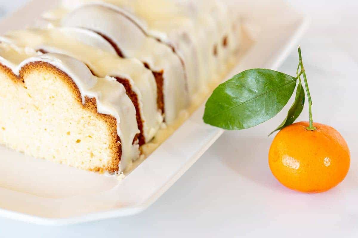 An orange pound cake covered in frosting on a white platter, oranges in the background.