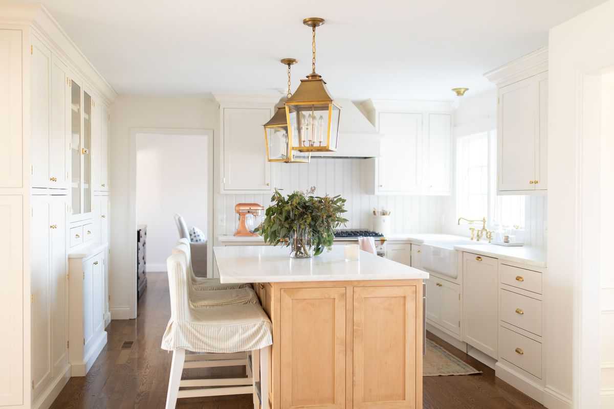 A white kitchen with a wooden island, vase of fresh eucalyptus on the island under lanterns.