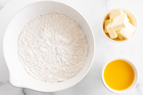 A bowl of flour beside smaller bowls containing cubed butter and melted butter, set against a marble background for making orange pound cake.