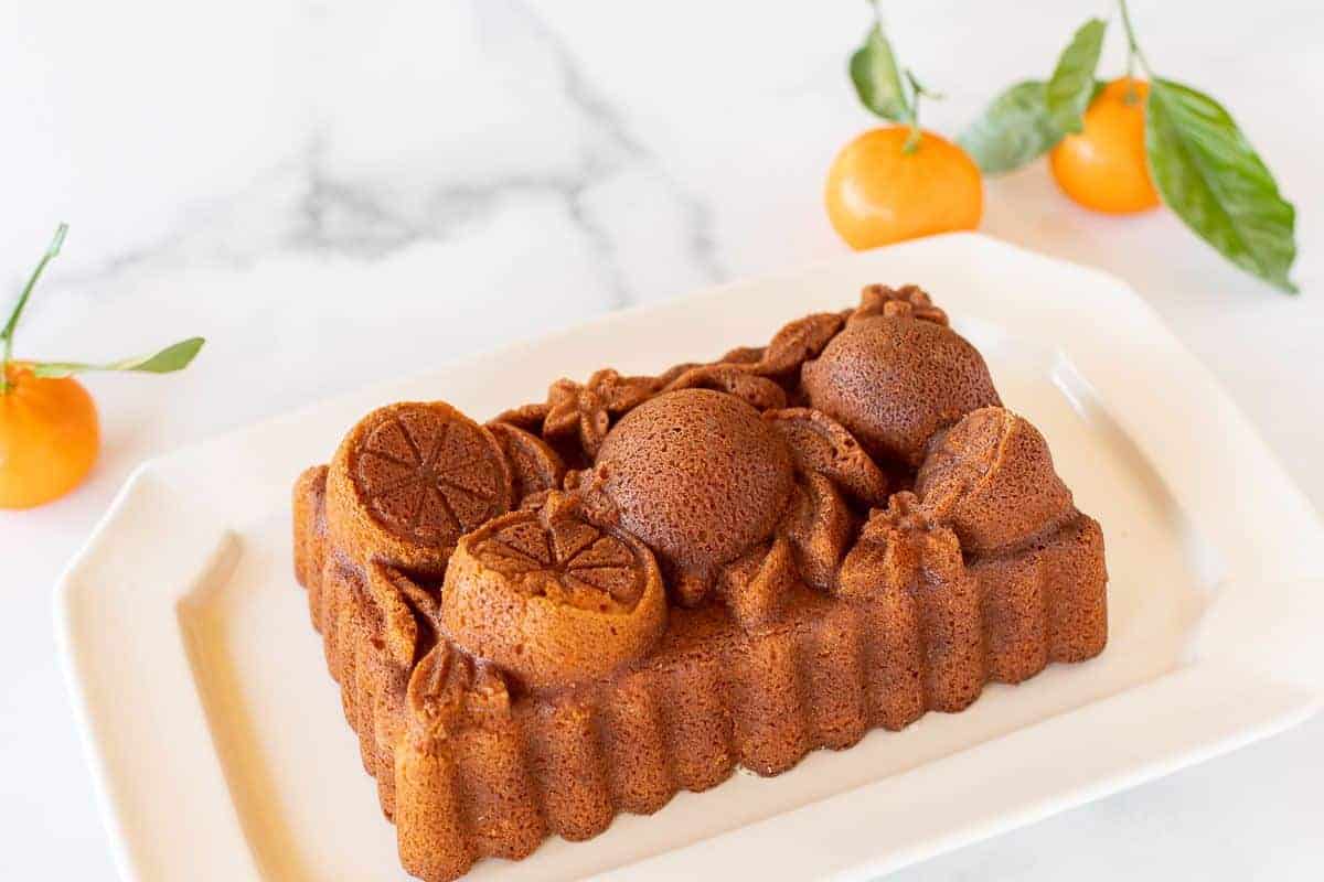 An orange pound cake from a citrus loaf pan, on a white platter. Oranges in the background.