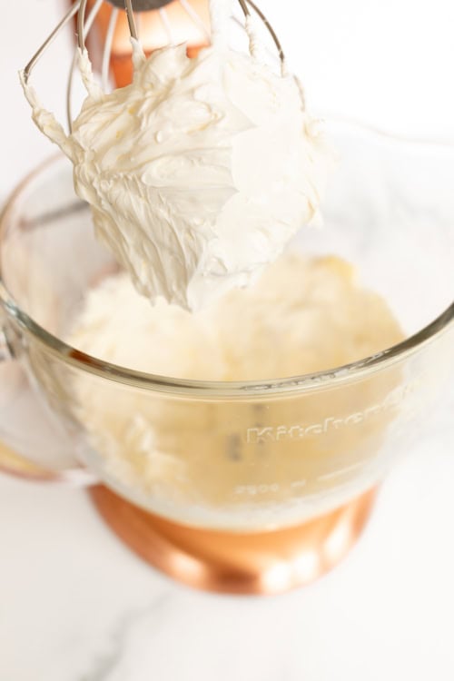 Electric mixer whisk lifting out of a glass bowl with creamy orange pound cake batter, emphasizing the thick texture clinging to the whisk.