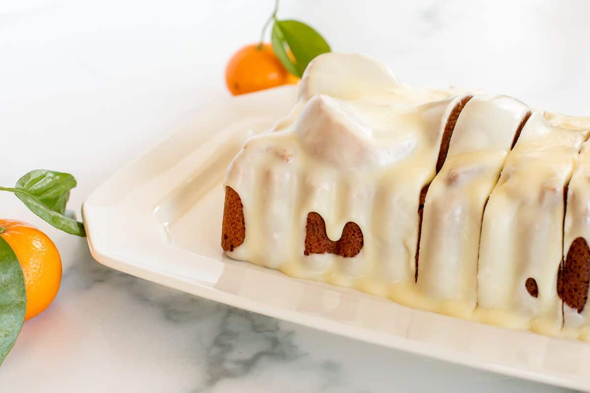 An orange pound cake covered in frosting on a white platter, oranges in the background.