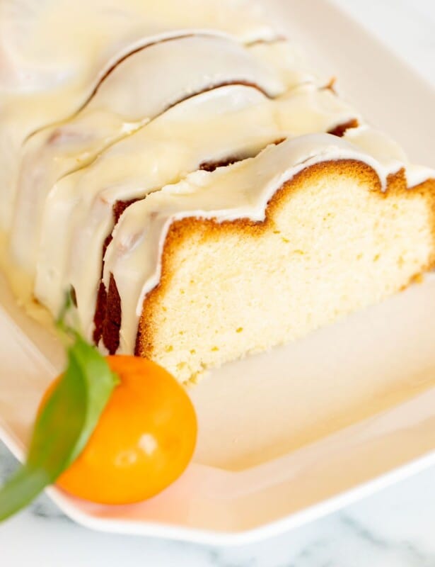 A sliced orange pound cake covered in frosting on a white platter, oranges in the background.