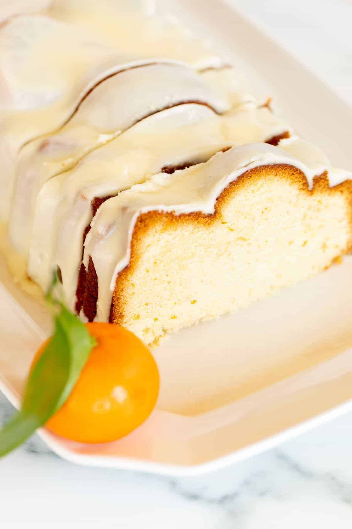 A sliced orange pound cake covered in frosting on a white platter, oranges in the background.