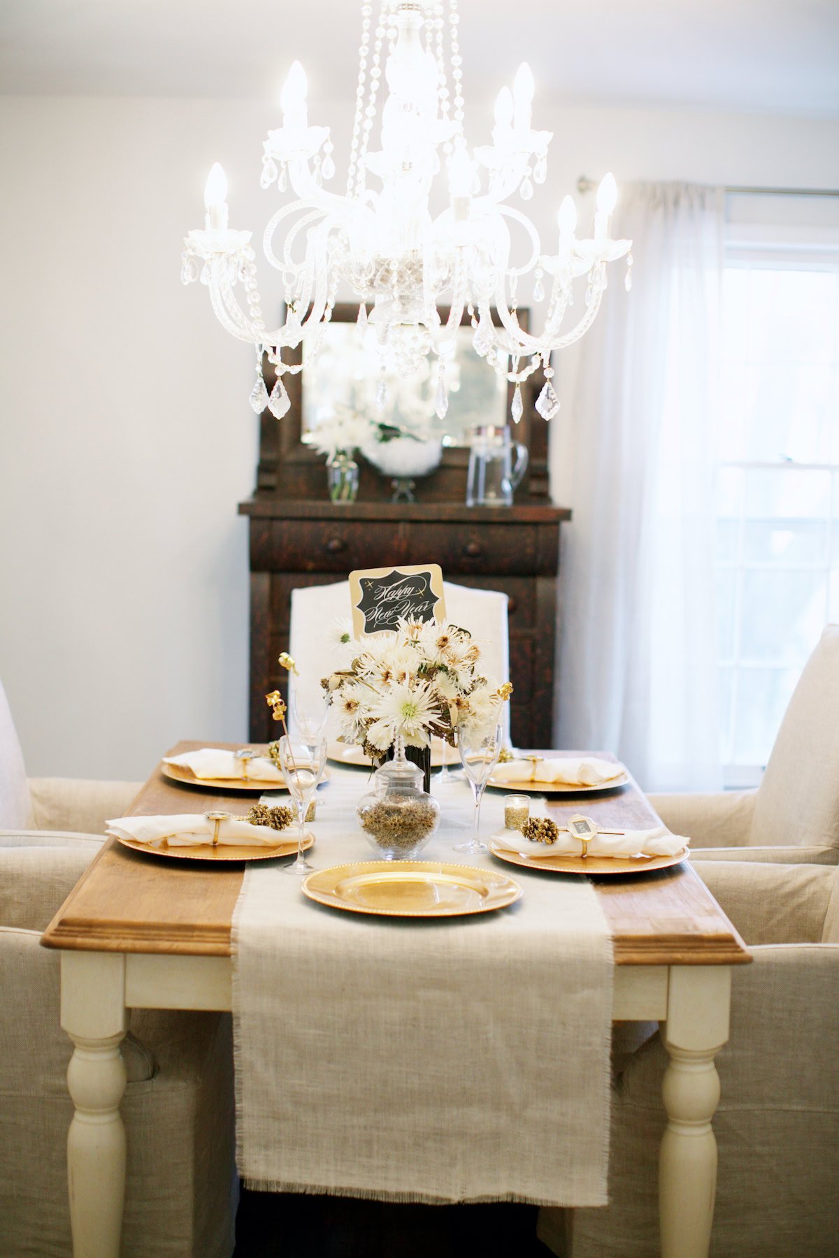 A dining table set for four with gold-trimmed plates, glasses, and a white floral centerpiece beneath a chandelier in a bright room painted in Sherwin Williams Accessible Beige, with a window and cabinet in the background.