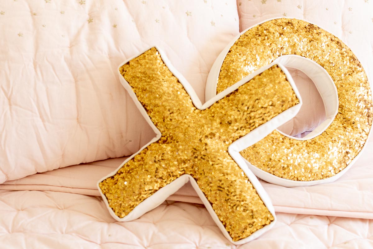 A gold sequined XO pillow set on a blush pink bed.