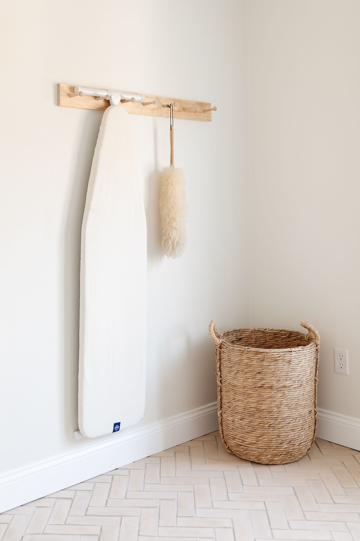 A peg rail on a white wall holding an ironing board and feather duster.