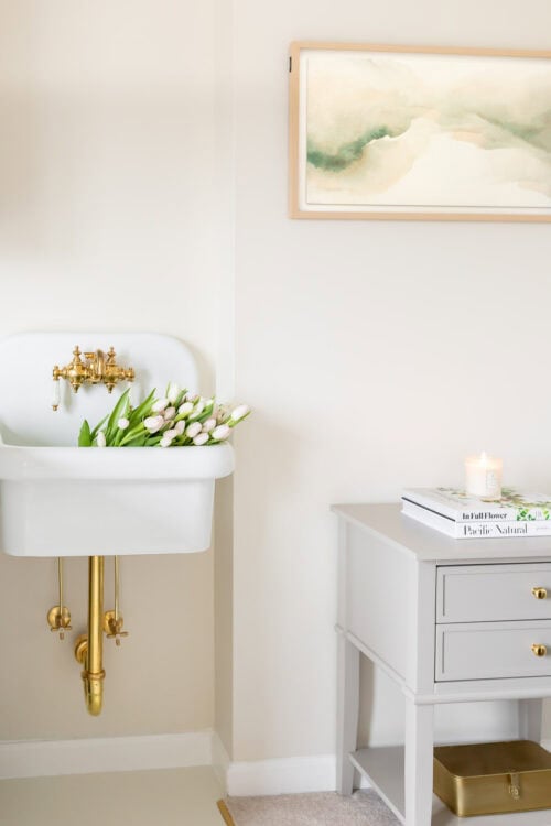 A white wall-mounted sink with brass fixtures holds a bouquet of tulips; next to it, a gray table with books, a lit candle, and a gold tray sits below a Samsung Frame TV displaying watercolor art.