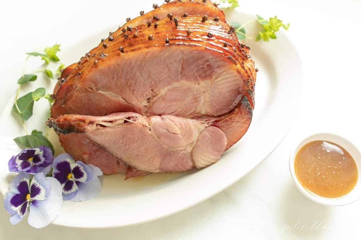 A honey ham studded with cloves on a white platter, garnished with fresh flowers for Easter
