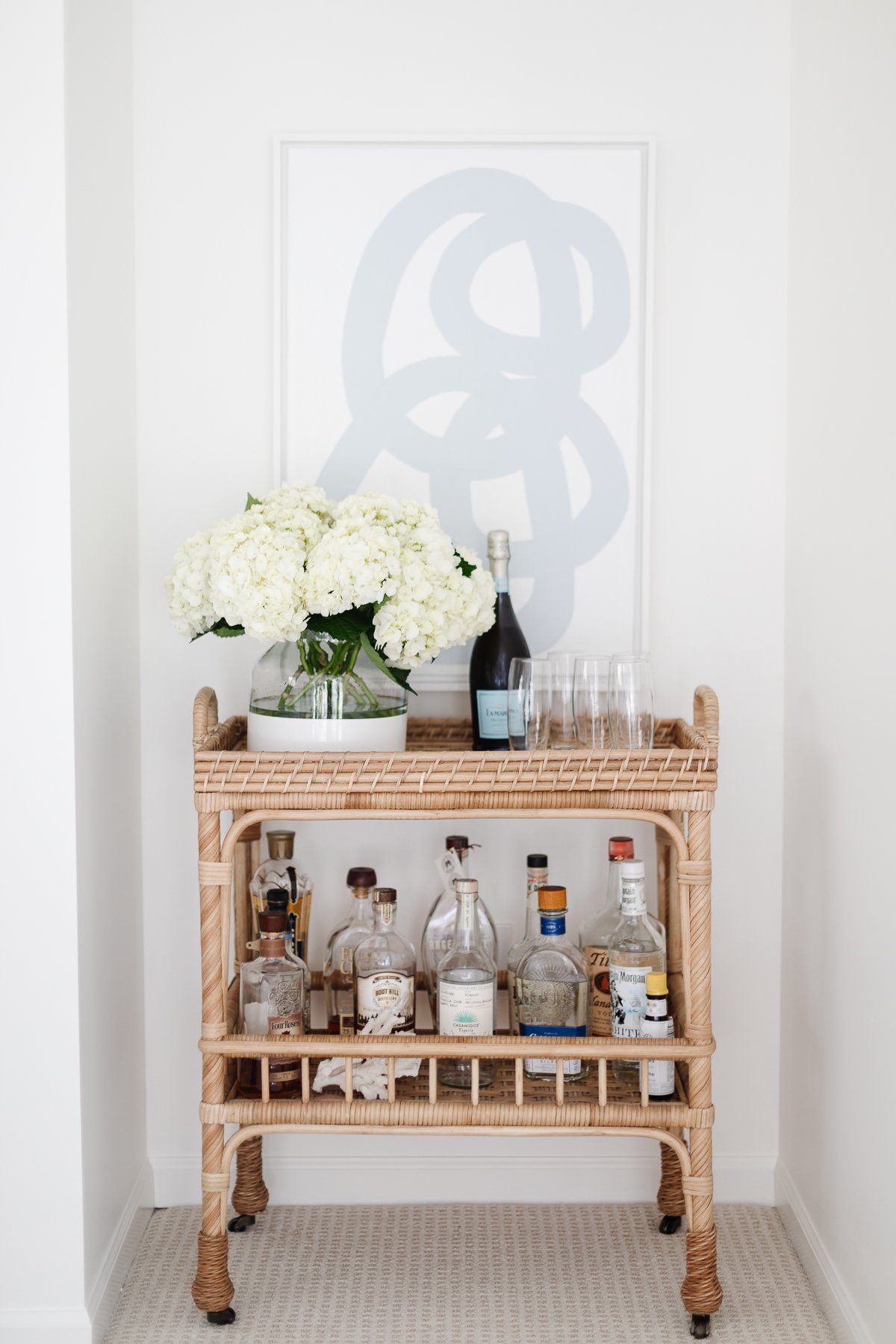A rattan bar cart with fresh flowers, bottles of alcohol and modern blue and white art in the background.