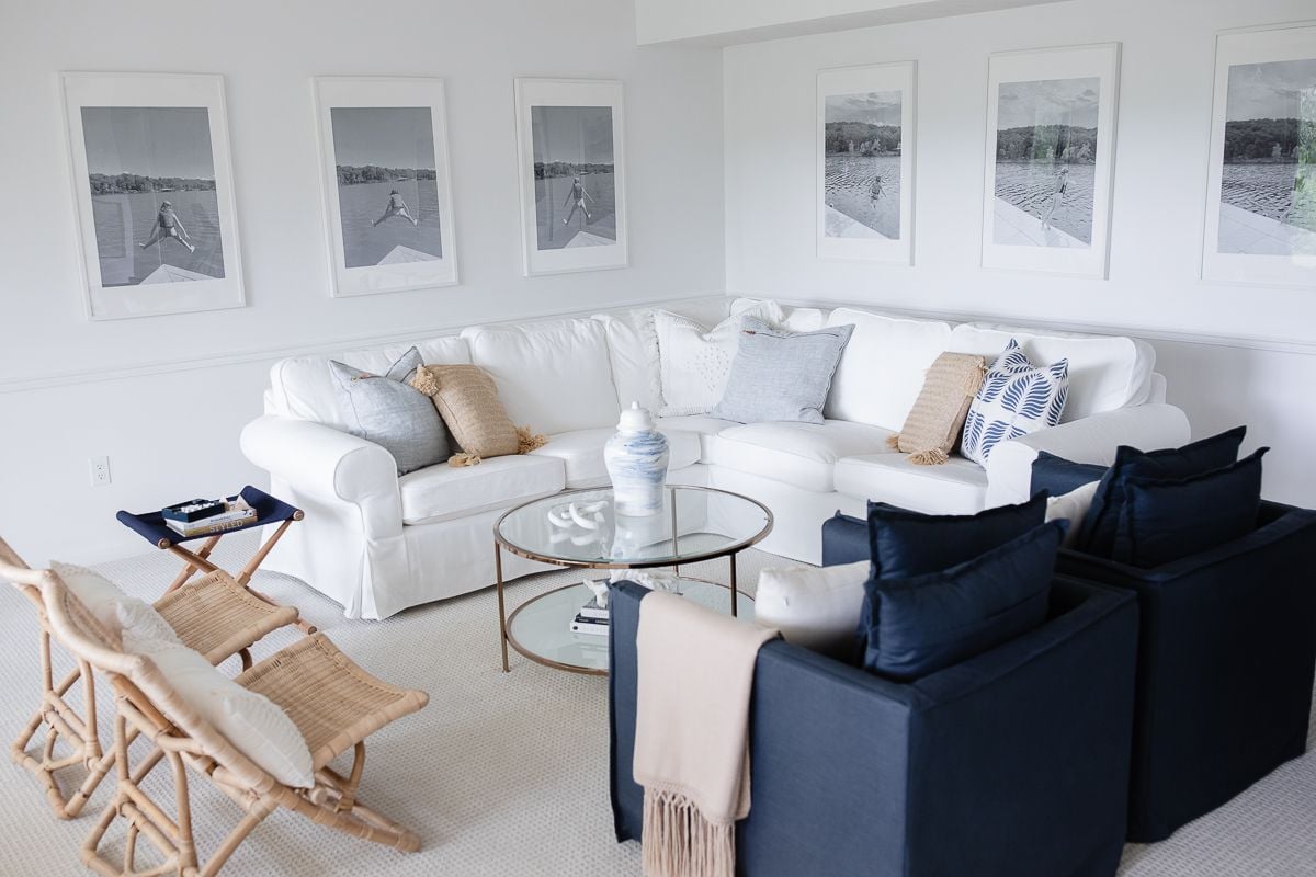 A white coastal modern living room with a white sectional, rattan folding chairs, and two navy upholstered chairs