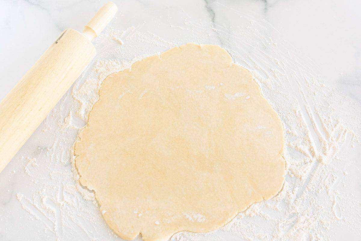 A homemade crust rolled out on a marble surface, wooden rolling pin to the side.
