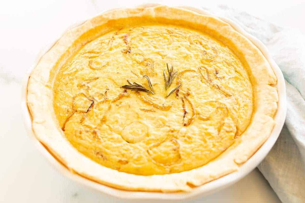 A fully baked quiche in a white pie dish on a marble surface, topped with fresh herbs.