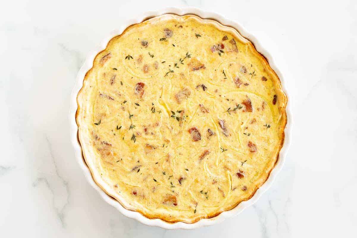 A whole quiche in a white pie dish on a white marble surface.