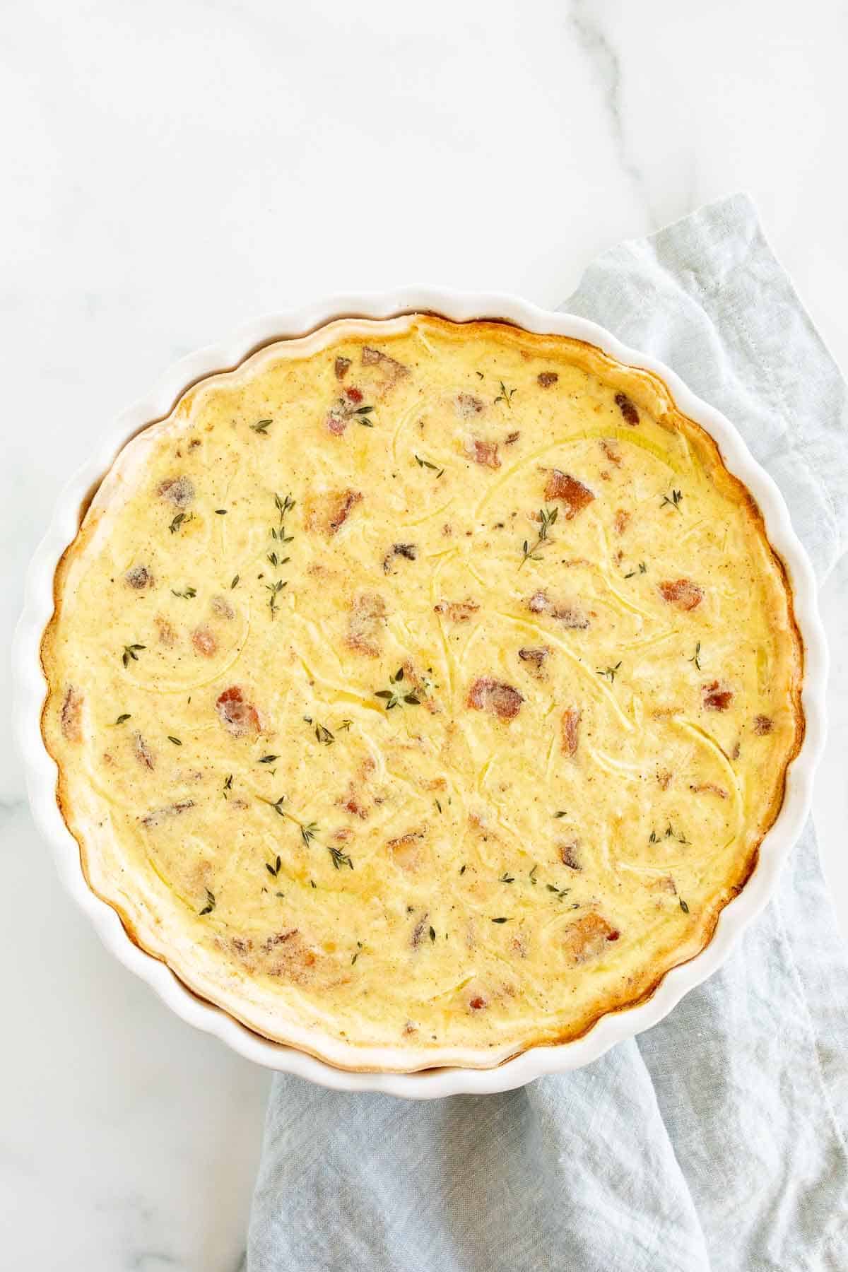 A homemade quiche in a white pie pan on a marble surface, blue towel to the side.